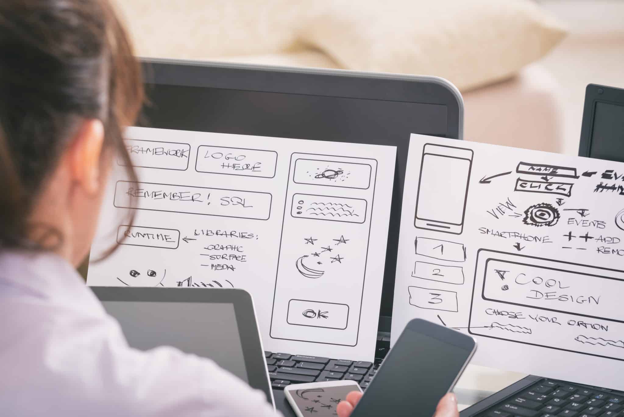 Ein Webdesigner überprüft handgezeichnete Layout-Skizzen für eine Website auf seinem Laptop.