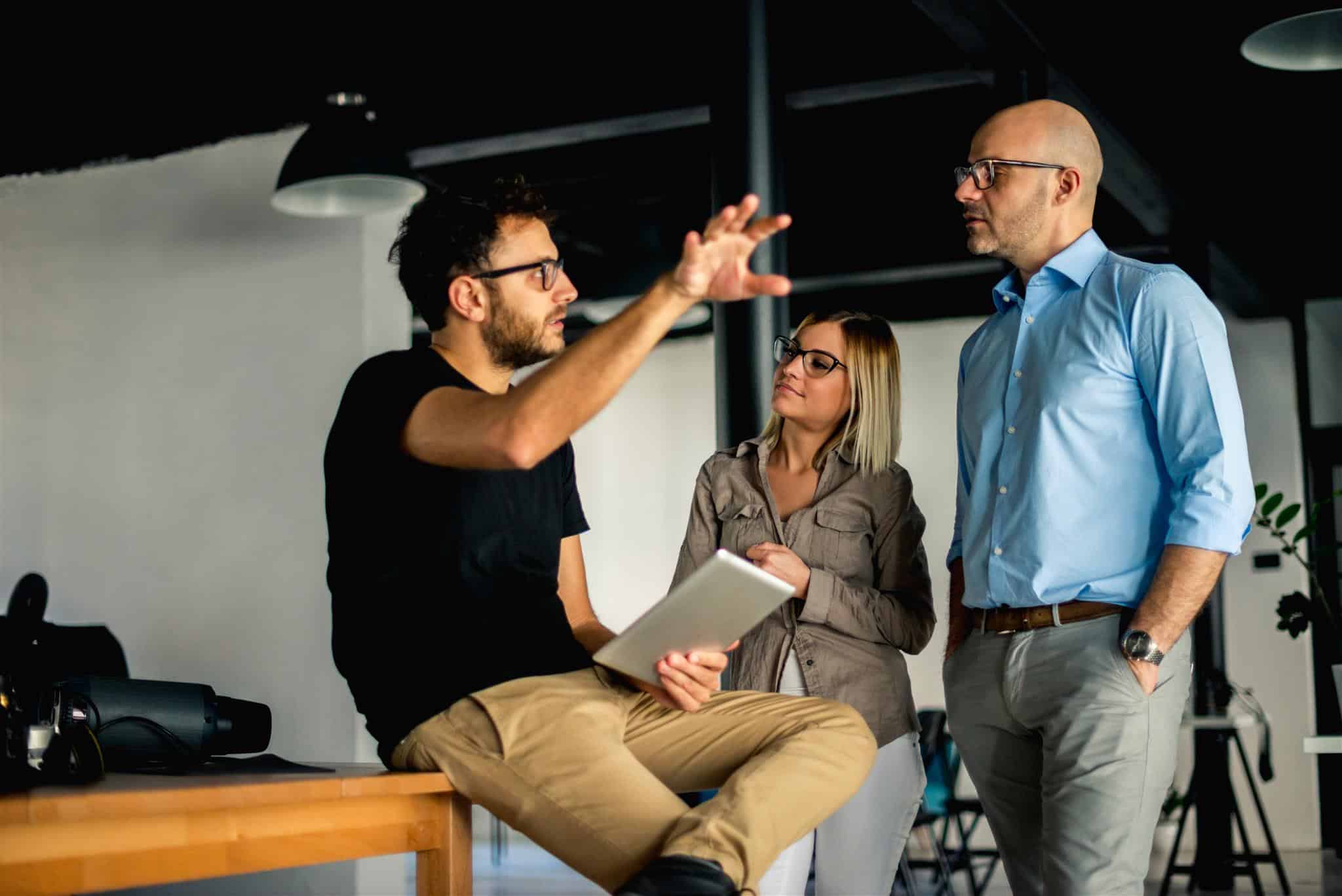 Team einer Digitalagentur bespricht ein Konzept im Büro: Ein Mann mit Tablet erklärt gestikulierend eine Idee, während zwei Kollegen aufmerksam zuhören.